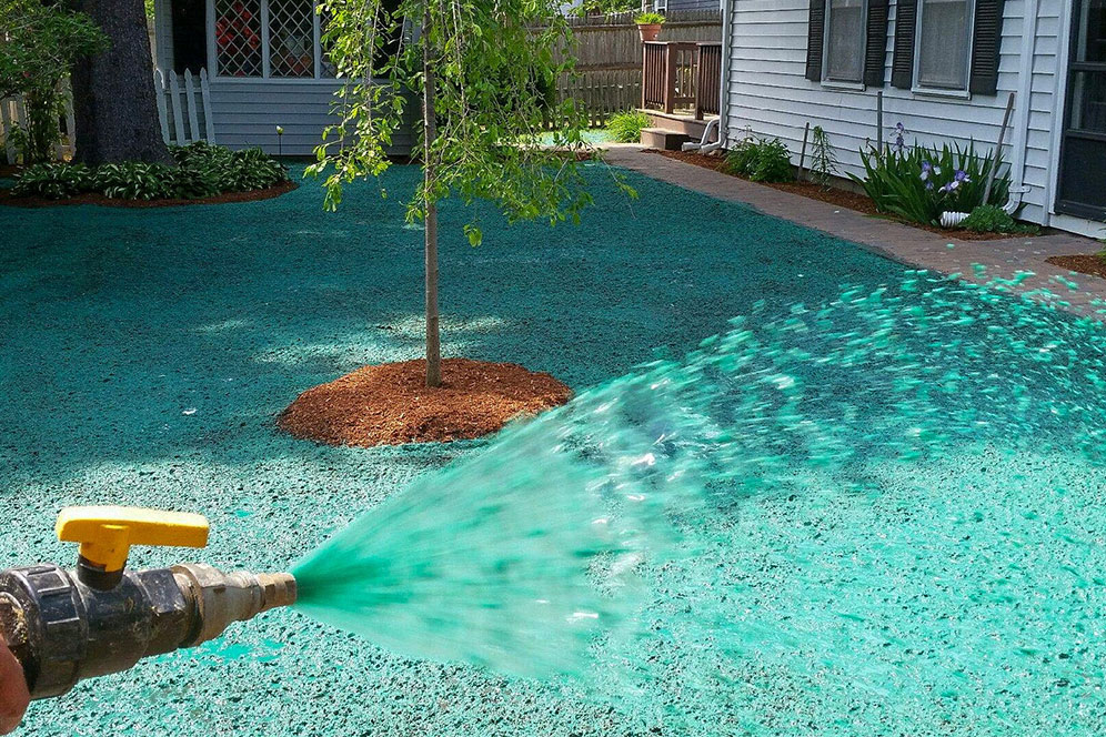 A person applies a green spray to a driveway, wearing gloves and aiming the nozzle carefully at the surface.