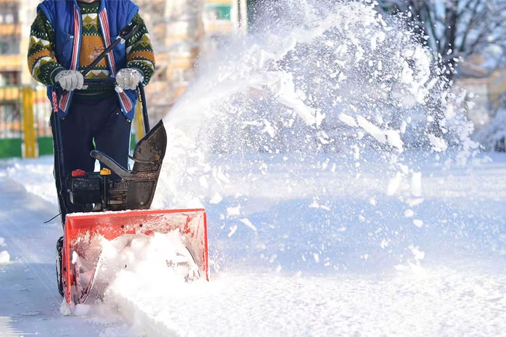 A man operates a snow blower, clearing a path through freshly fallen snow in a residential area.
