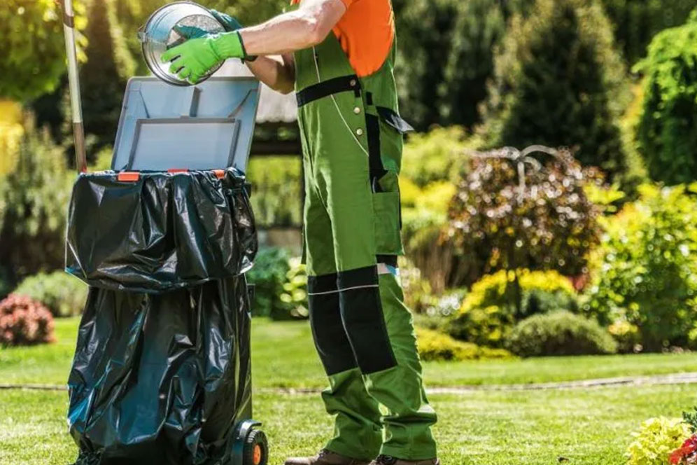 A man in overalls and green work clothes stands holding a trash can, ready to collect waste.